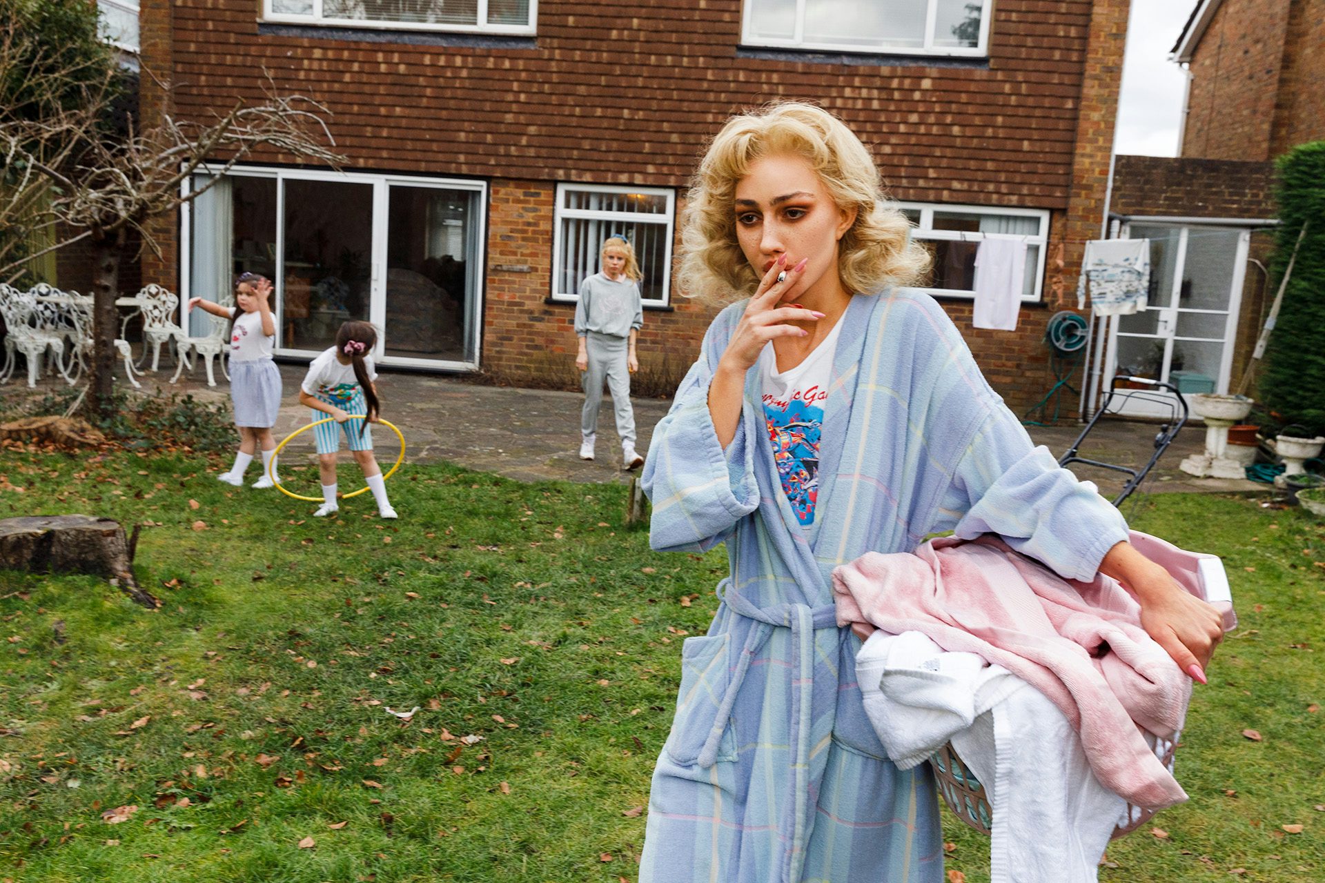Photo from Julie Bullard by Nadia Lee Cohen and Martin Parr of a young blonde woman in a bathrobe holding laundry in a garden smoking a cigarette with children in the background