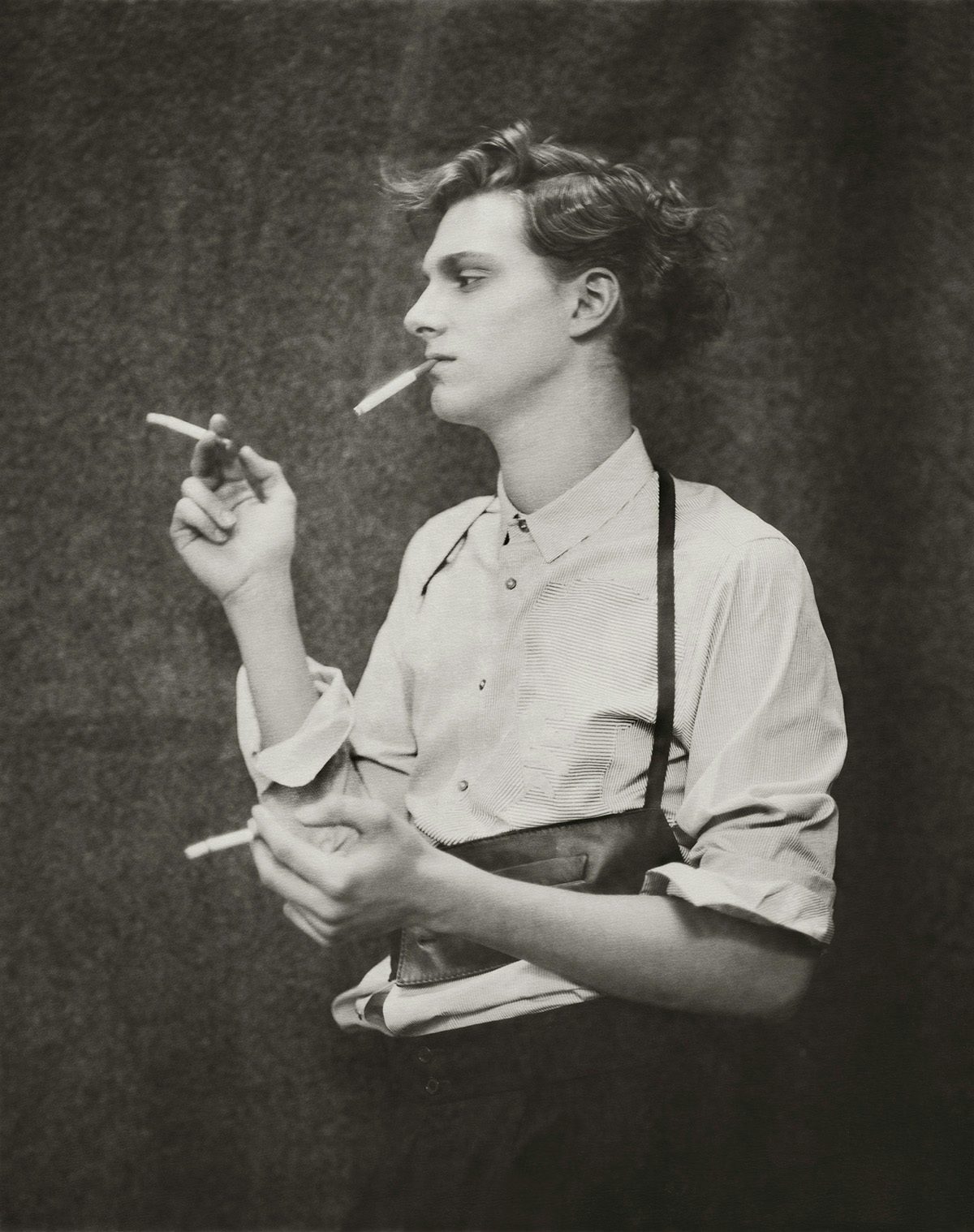 Black and white photo by Paolo Roversi of a model white short wavy hair and wearing a light buttoned shirt with a wide leathery belt, smoking a cigarette and holding more in each hand