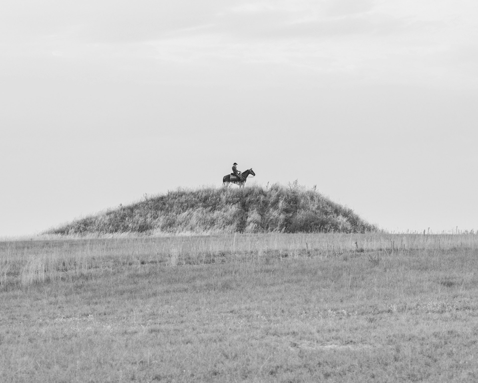 Black and white photo by Karol Szymkowiak showing a person at the top of a small mound riding on the back of a horse in the distance