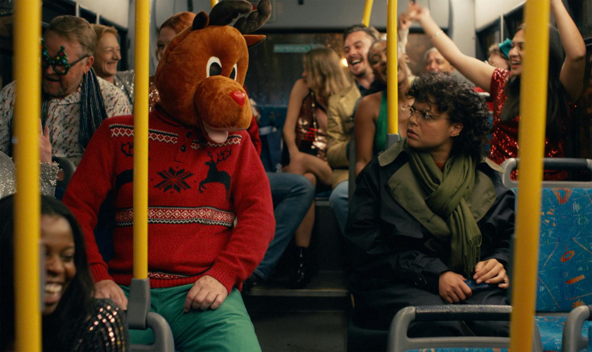 Still image of a person sat on the bus with a reindeer head on facing another person sat across the aisle looking confused