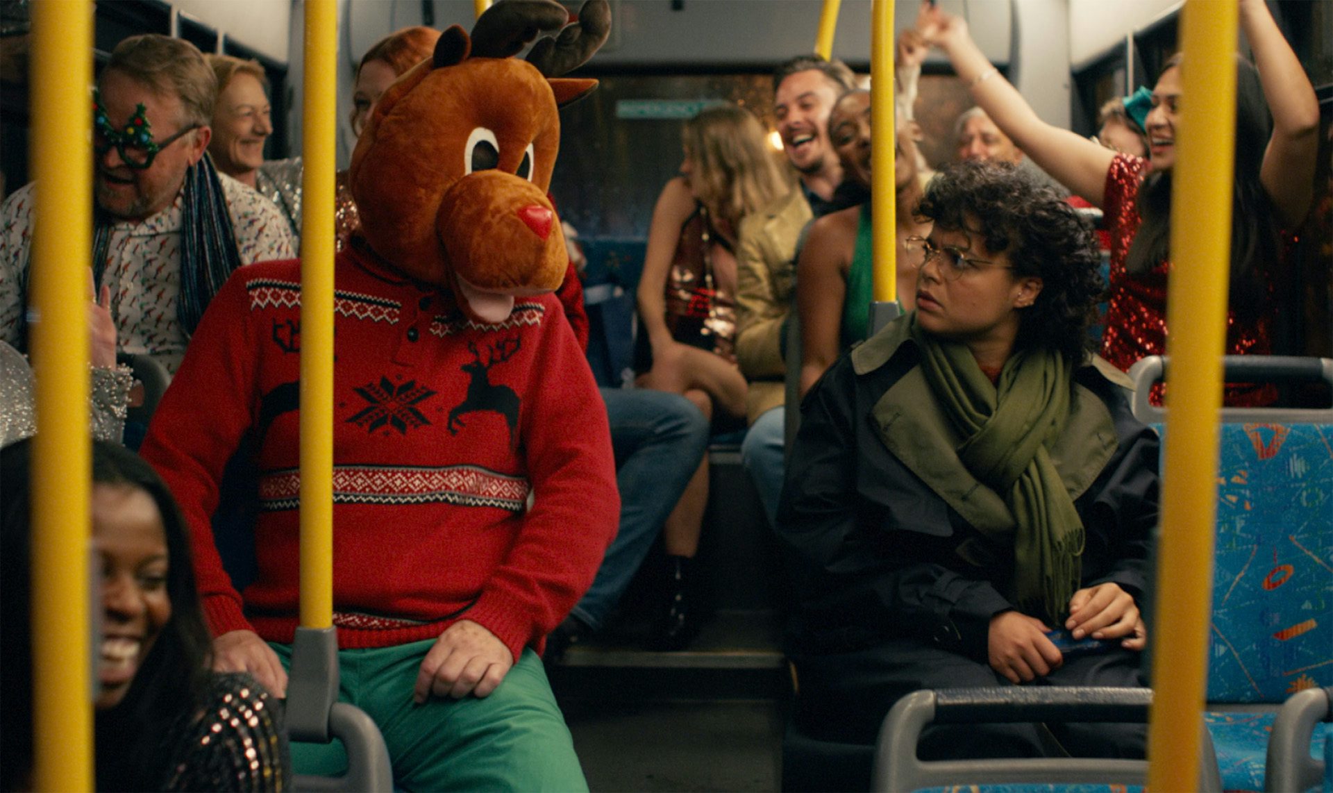 Still image of a person sat on the bus with a reindeer head on facing another person sat across the aisle looking confused