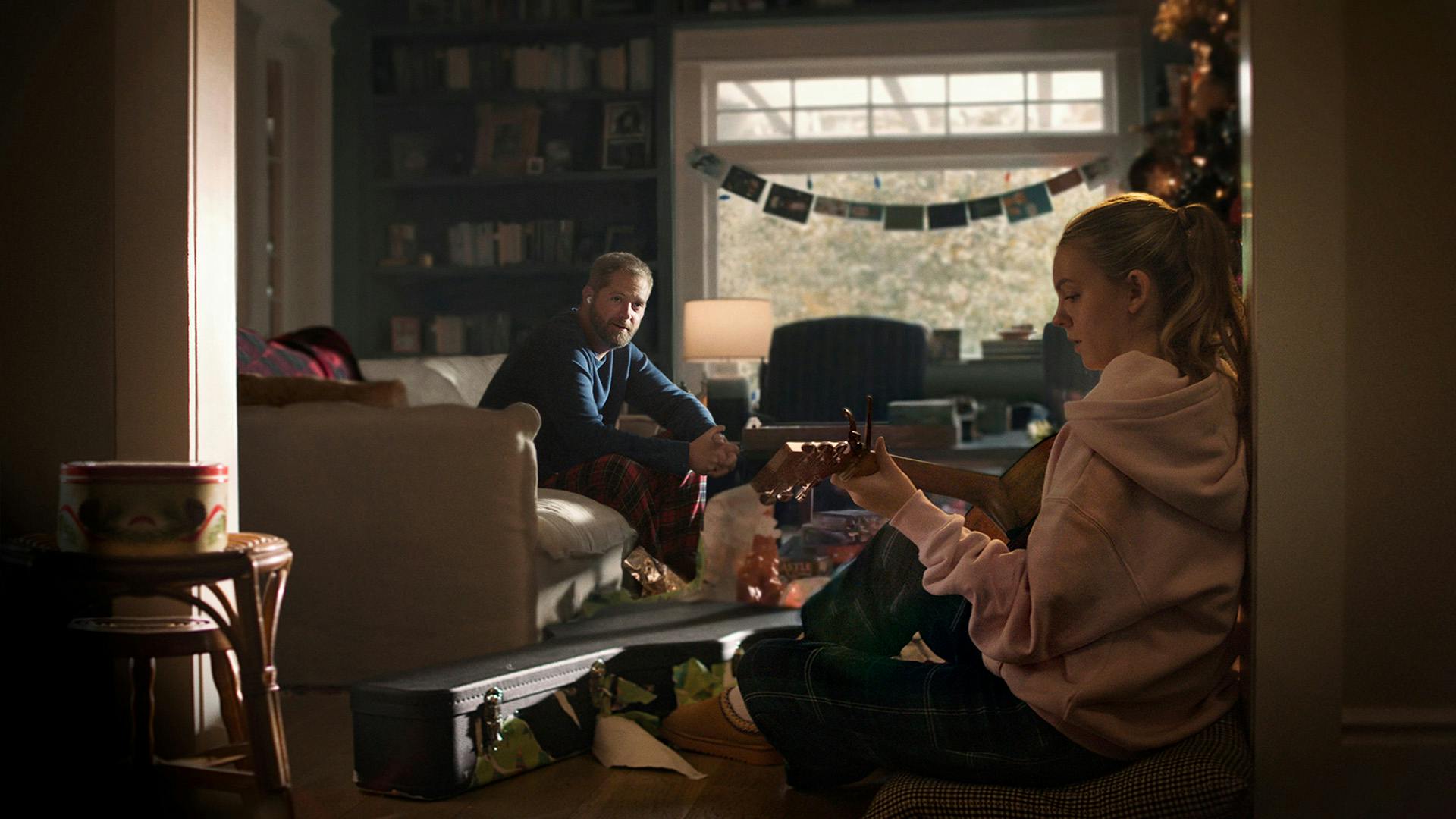 Still from Apple's Heartstrings ad showing a man sat on a sofa in a living decorated for Christmas watching his daughter play guitar in the foreground