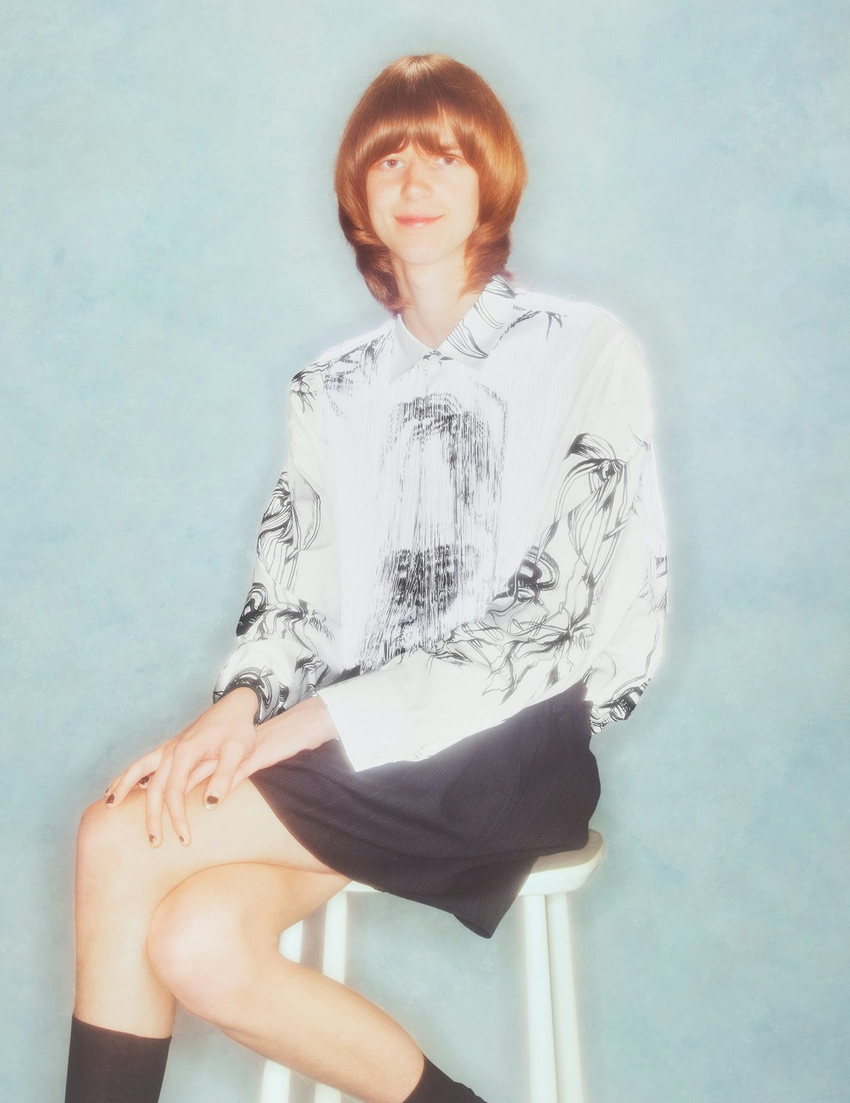 Photo by Jakub Gessler of a person with a bowl cut wearing a white patterned shirt and black skirt and sat on a stool against a studio backdrop