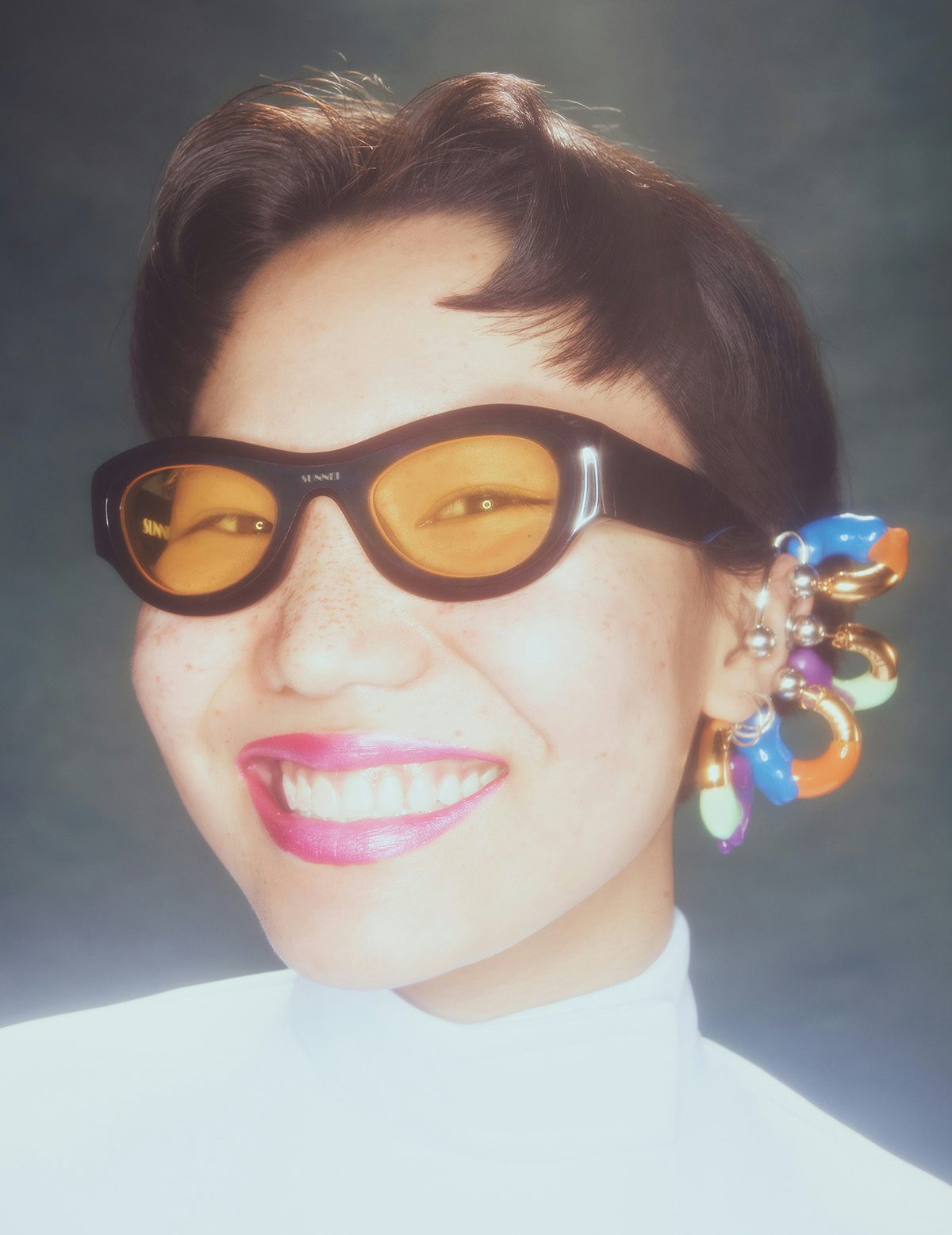 Portrait photo by Jakub Gessler of a person with short hair wearing black oval sunglasses and a stack of colourful hooped earrings on their ear