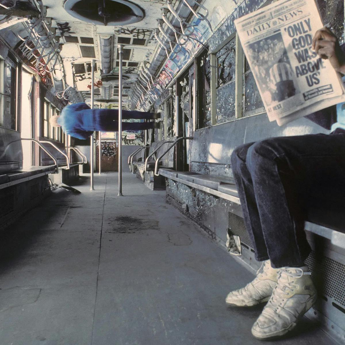 Album art for Only God Was Above Us by Vampire Weekend shot inside an abandoned subway that is tilted on its side, showing a person sitting on a seat reading a newspaper with the a headline the same as the album name, and another person in the background appearing to be walking on the side of the carriage