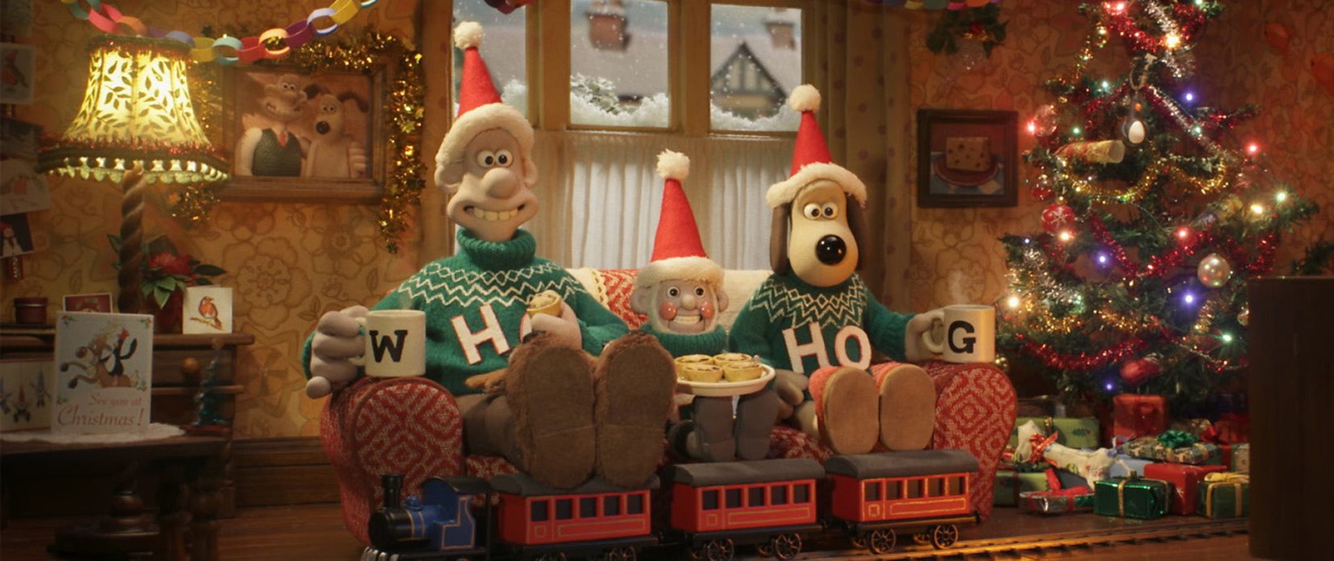 Still from a new BBC One ident showing animated characters Wallace and Gromit sat on a sofa in a Christmas decorated living room with a gnome in between them, all wearing red Christmas hats