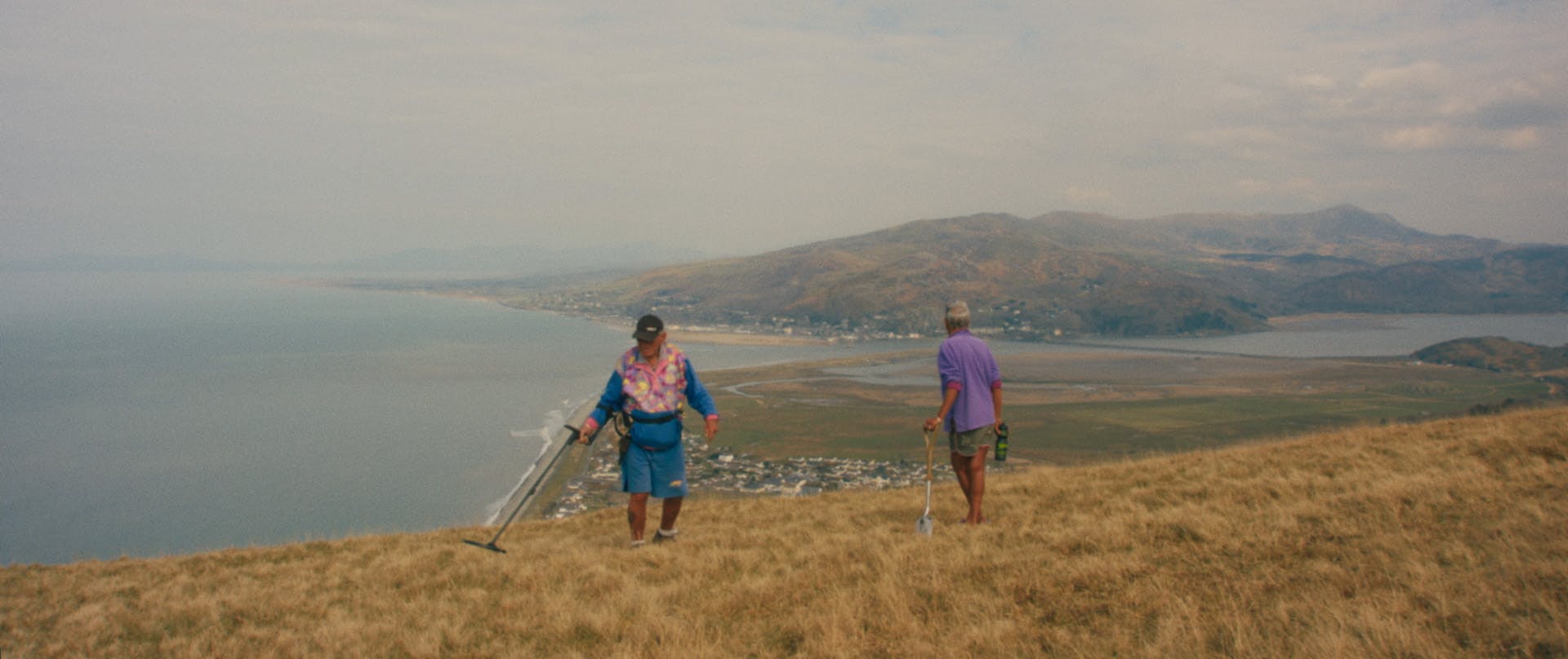 Two people standing on a coastal hillside holding devices resembling metal detectors