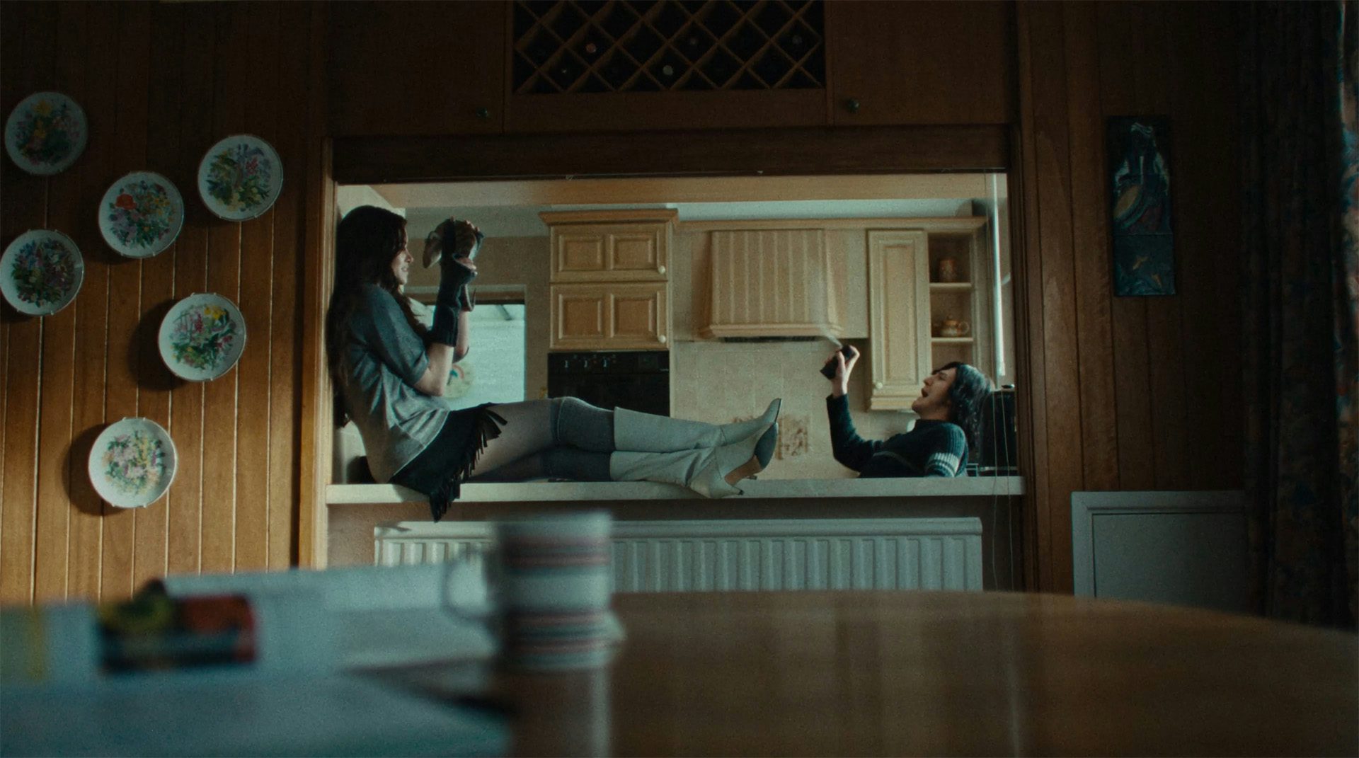 Still from the Fontaines DC music video, It's Amazing To Be Young, showing a person sat on top of a kitchen counter holding a snake with another person blowing an aerosol can in the kitchen in the background