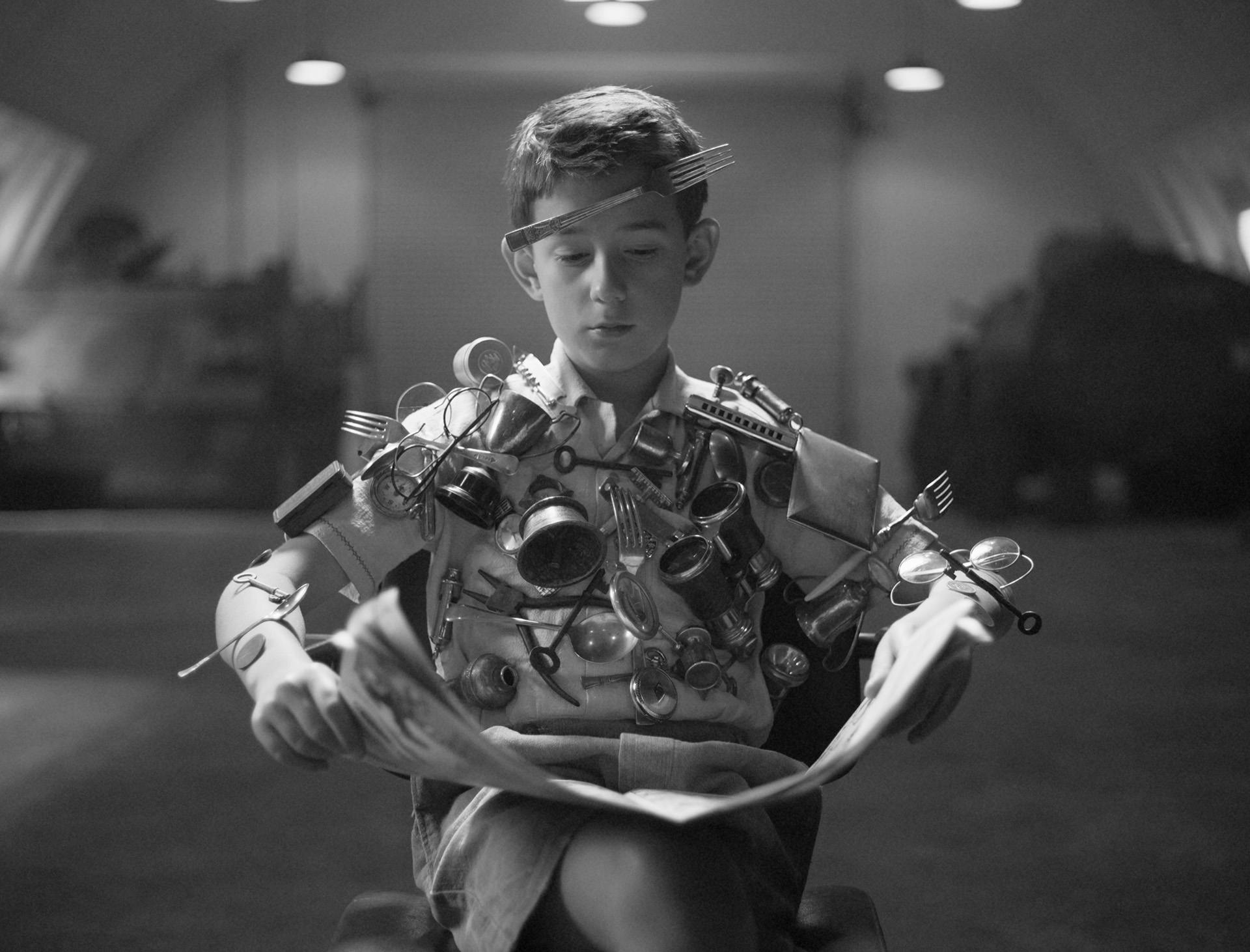 Black and white still from a Telstra short by Dougal Wilson showing a young boy sat on a chair reading a newspaper with metal objects fixed to his body and head