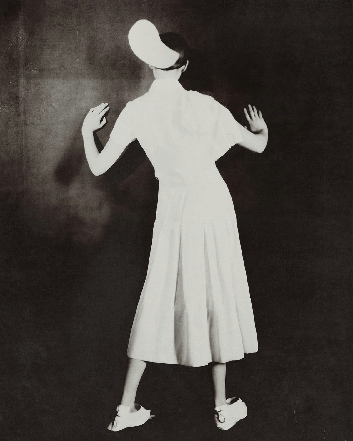 Black and white photo by Paolo Roversi of a person wearing a calf-length white dress and headpiece standing with their back to the camera