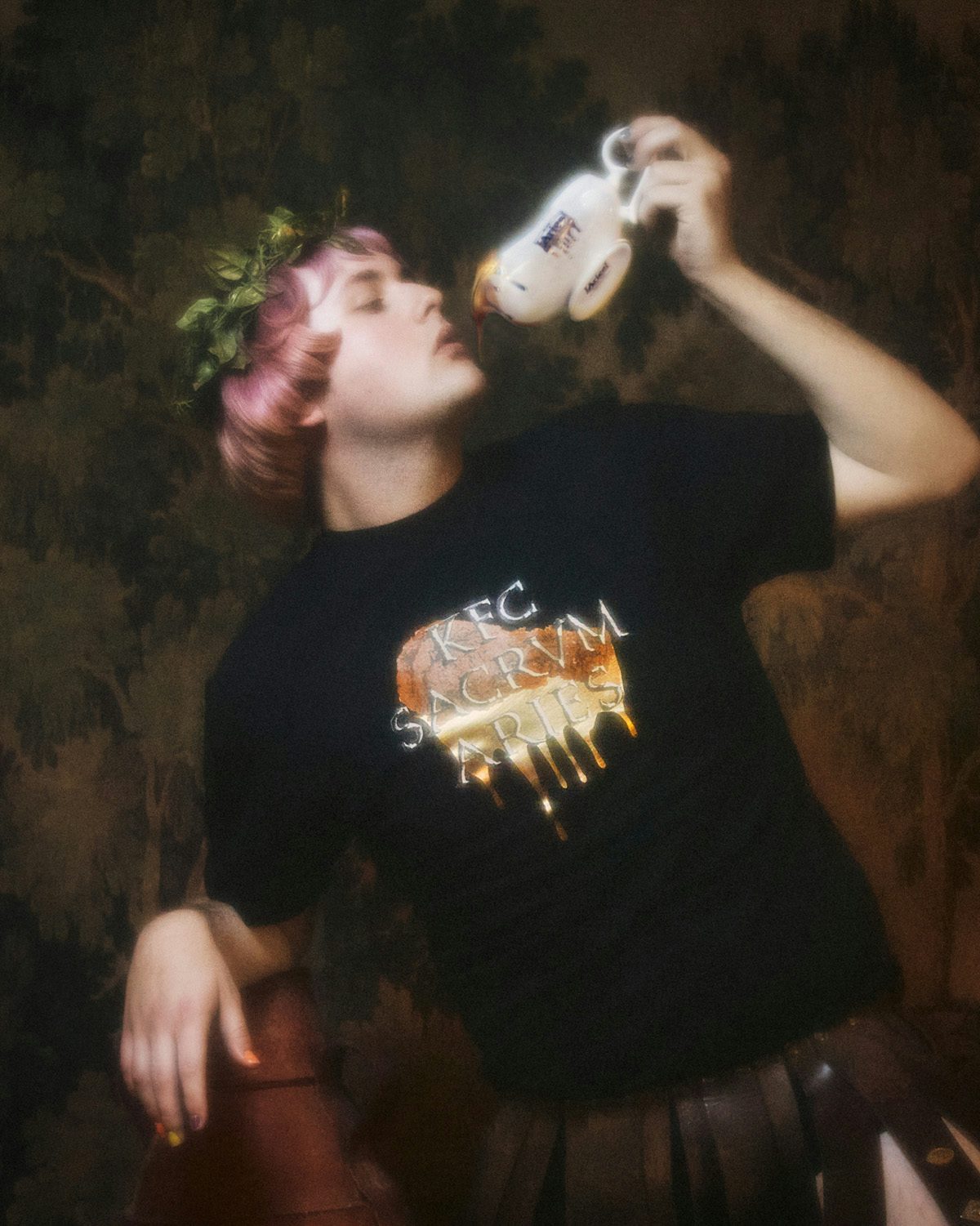 Soft focus image of a person with light pink hair wearing a laurel and a black t-shirt with the slogan 'KFC sacrum Aries' while holding a gravy boat towards their mouth