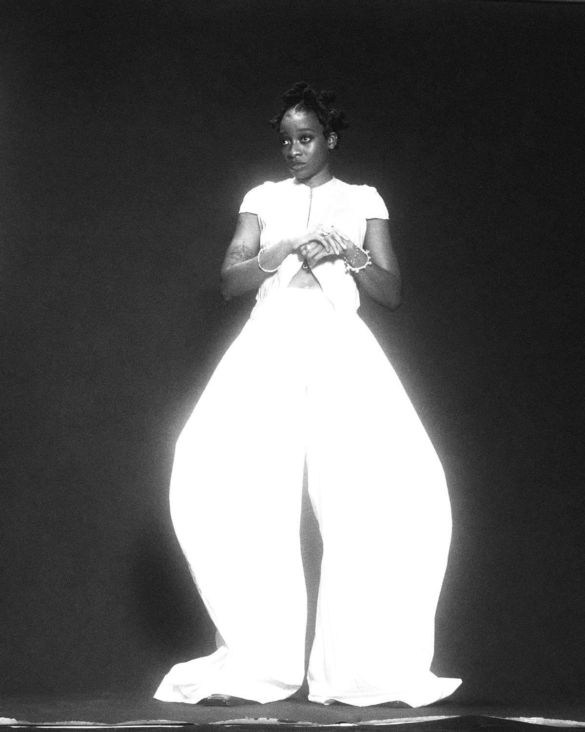 Black and white photo of Little Simz wearing a white top and ballooned wide-leg trousers by Thibaut Grevet